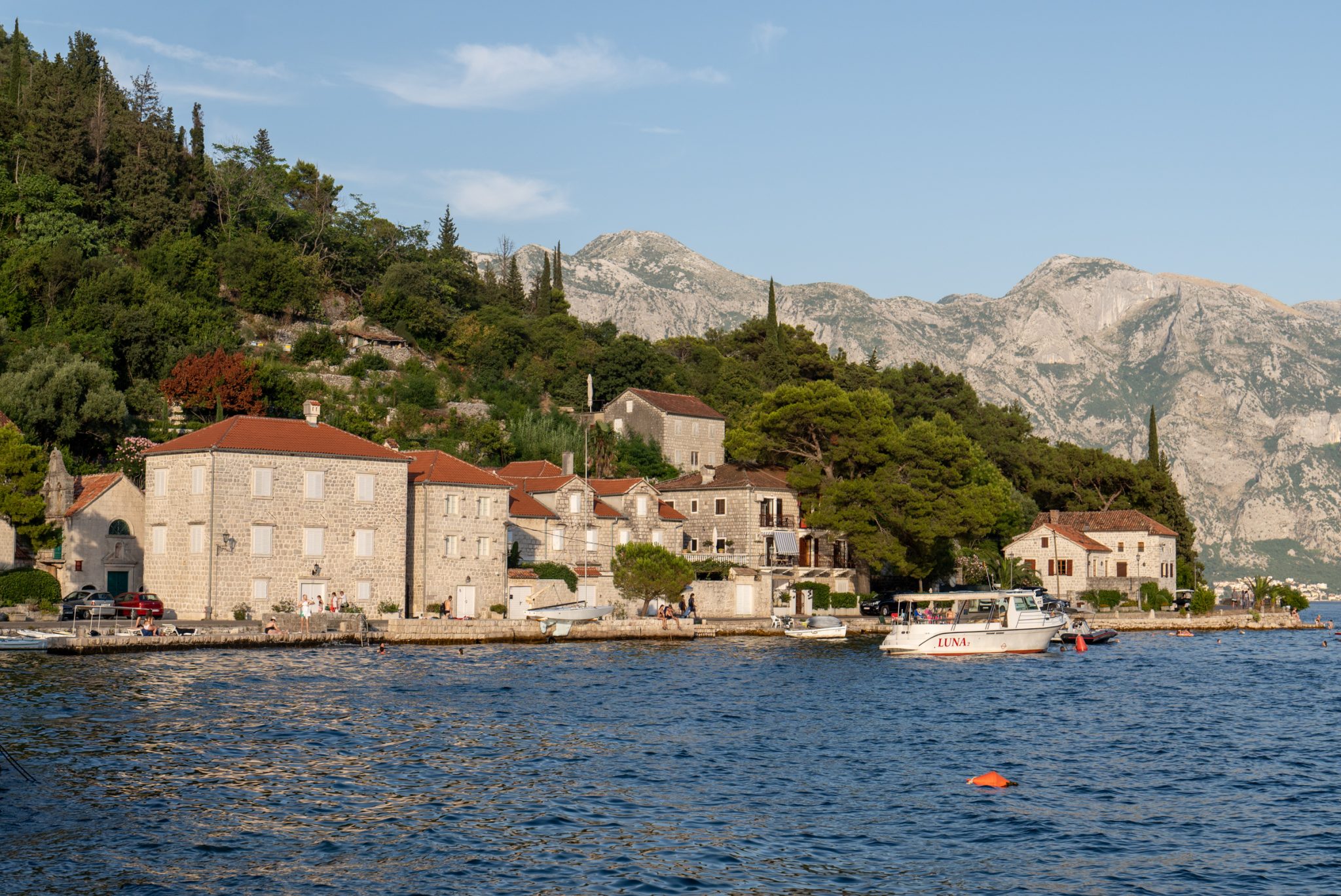 A tiny village of stone houses with orange roofs set against a forested hillside on the Bay of Kotor.