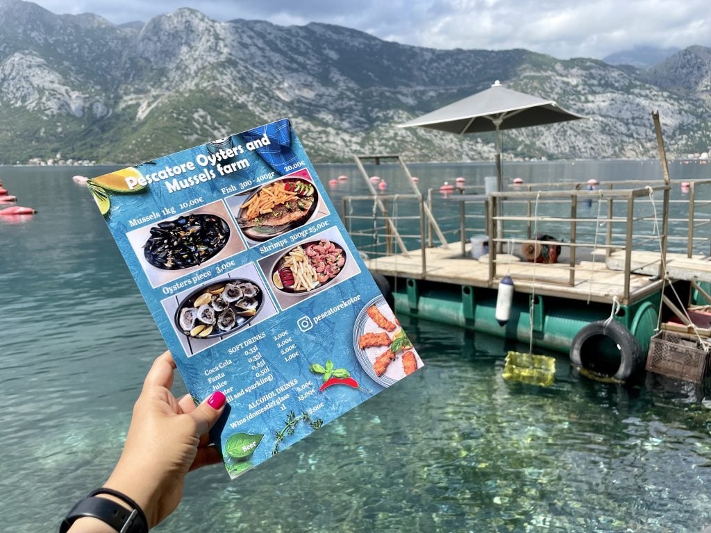 A menu of an oyster and mussels restaurant held over the clear green waters of the Bay of Kotor.