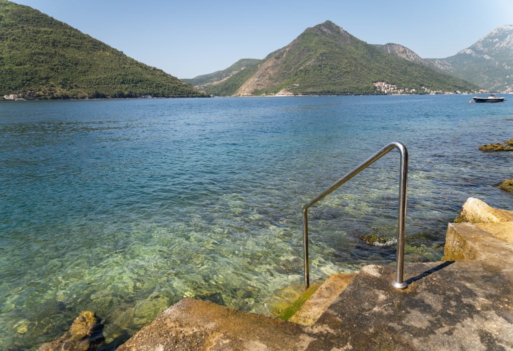 A metal pole on the edge of a concrete platform leading into the bright green-teal-blue Bay of Kotor, jagged green mountains in the distance.