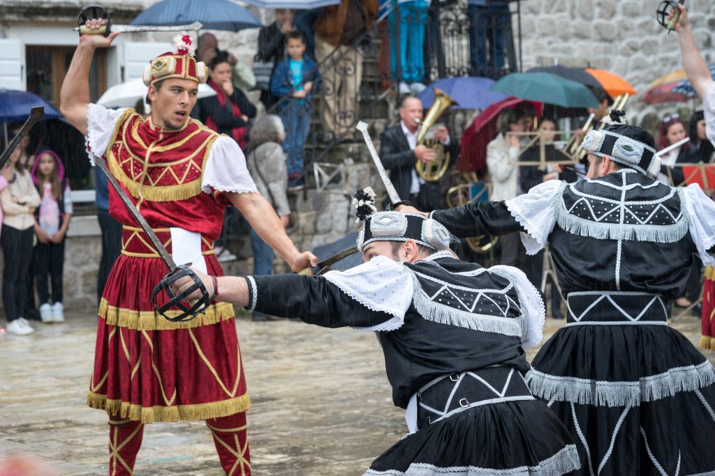 Men in traditional red and black satin gladiator outfits fighting with swords on a square in Perast.