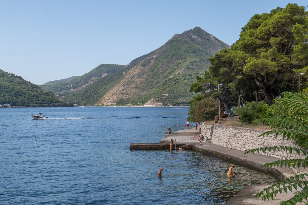 A long cement block on the bay, people swimming in the water, tall green mountains behind them.