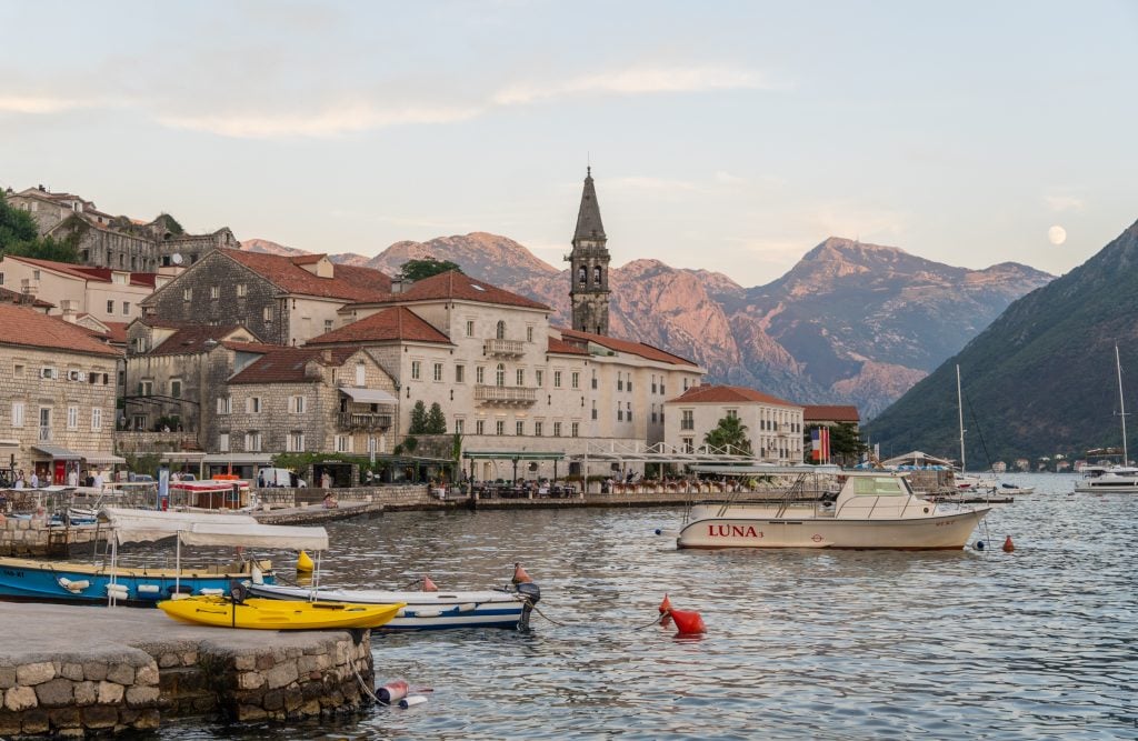 The quiet village of Perast at sunset, with lots of stone buildings with orange roofs and a pointy church tower, set against the mountainous landscape.