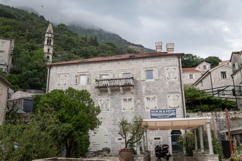 Stone buildings in Perast shot from below, surrounded by green trees, underneath low-hanging clouds.