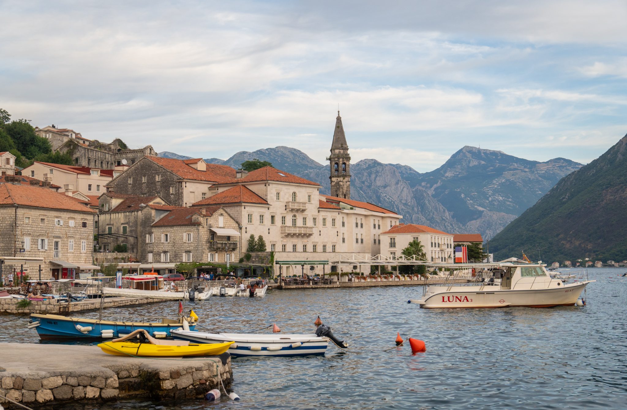 A view of Perast, a small town of small buildings with orange roofs, one bell tower sticking up, set on a peaceful calm bay and surrounded by mountains.