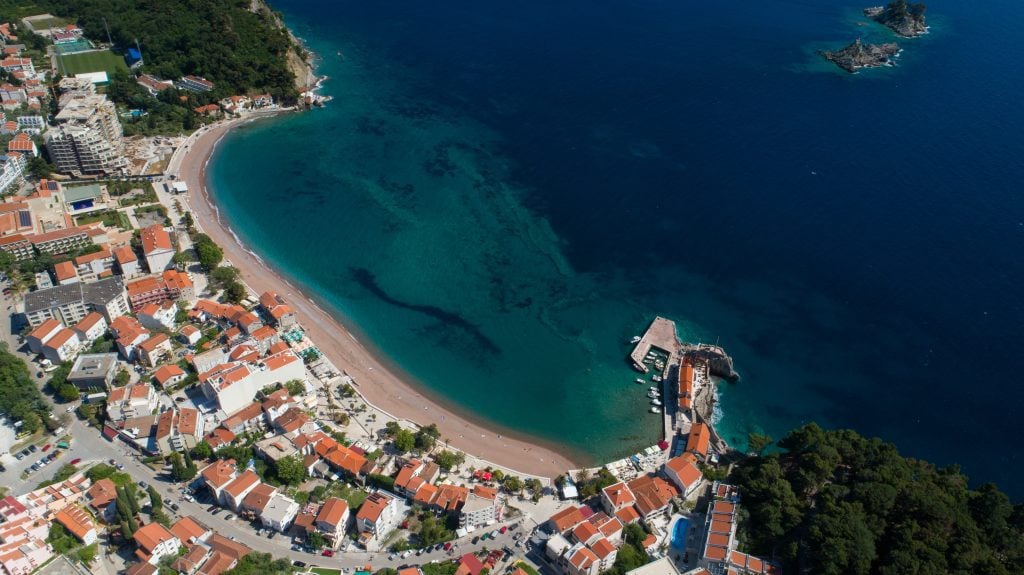 An aerial shot of the town of Petrovac, with its half moon of sandy beach surrounded by white and orange houses.