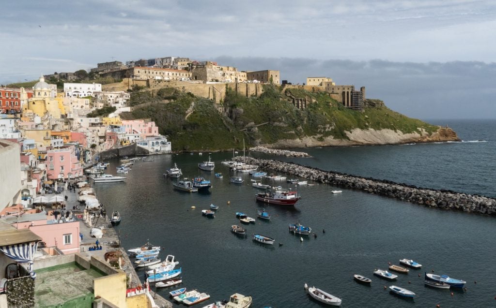 Procida's colorful marina, filled with pink, yellow, and orange houses in front of a bay filled with wooden rowboats.