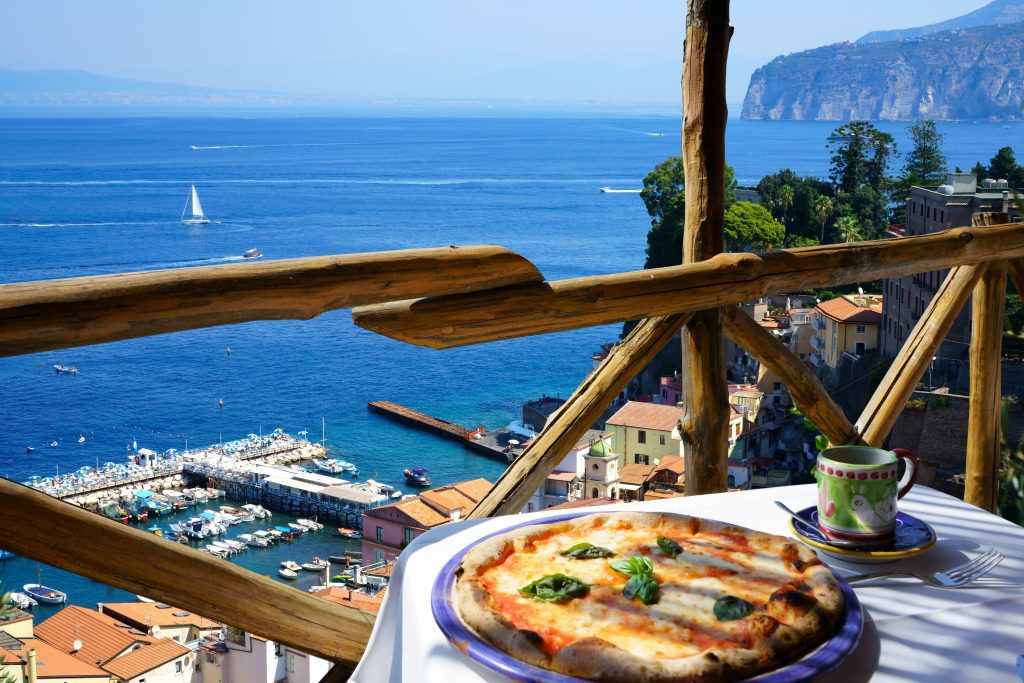 A pizza sitting on a table on a balcony overlooking the sea in Italy.