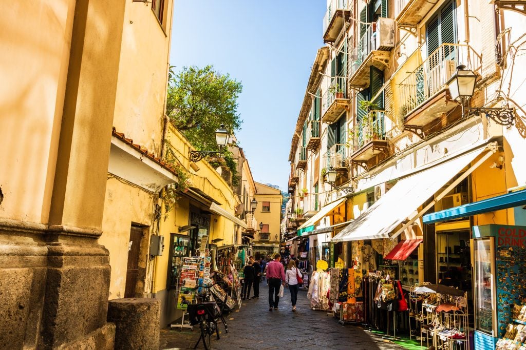 People walking down a busy Sorrento street with fruit stands and souvenir sellers underneath awnings.