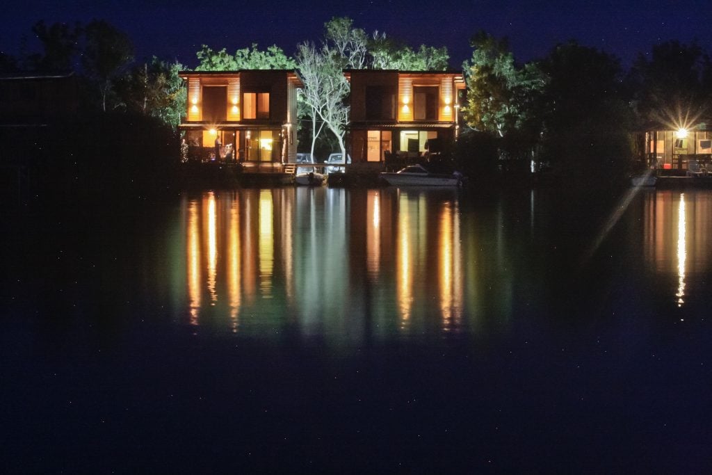 Two modern houses on the river lit up at night, reflecting in the inky water below.