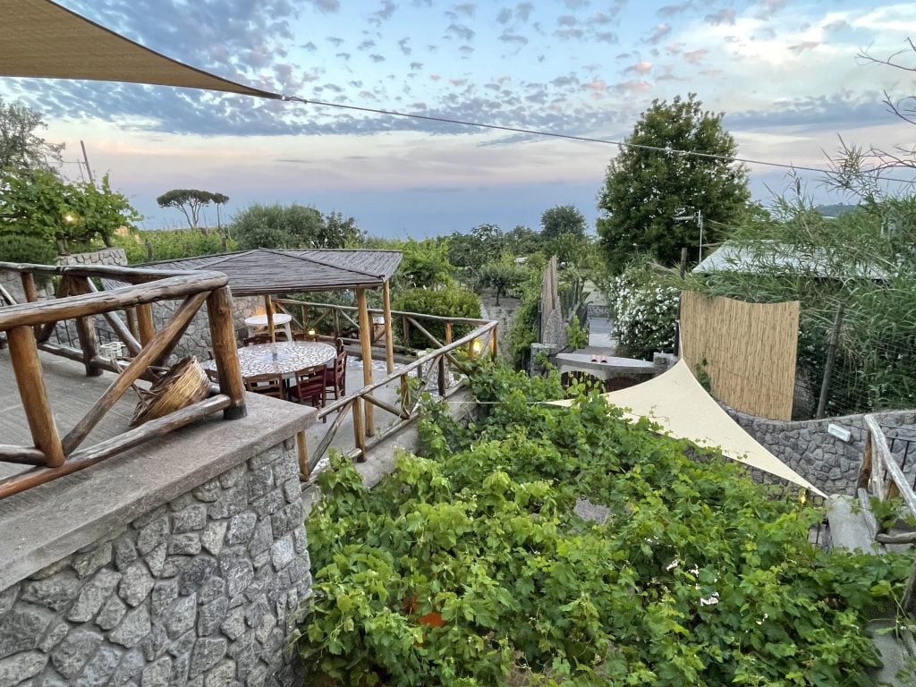 A gorgeous view of an agriturismo: outdoor seating area on terraces of a stone house, with lots of lush greenery below, and a sunset with purple spotted clouds across a pink and blue sky.