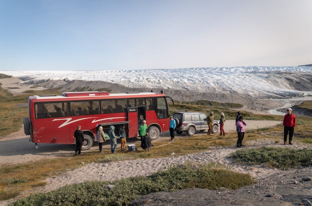 A big red bus parked in front of a green hill with a big glacier in the distance and lots of tour guests milling around.