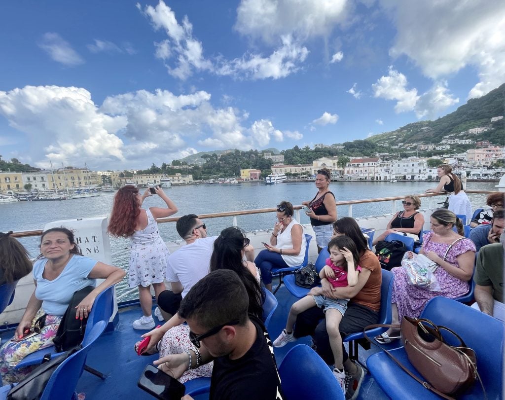 People standing on the balcony of the car ferry, posing for photos with the coastline of Ischia in the distance.