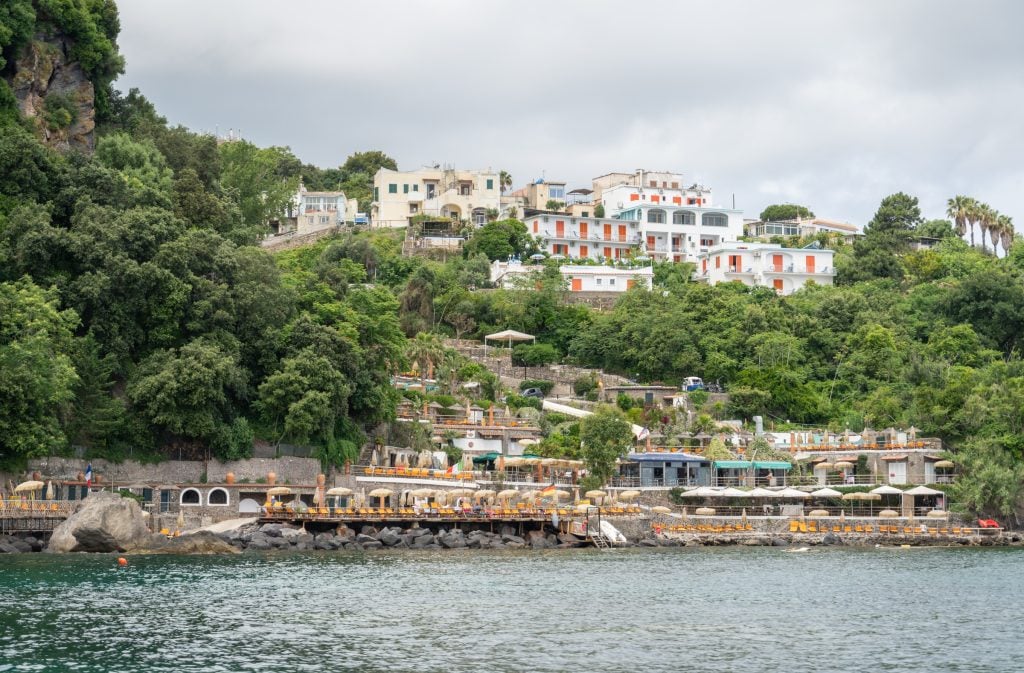 A whitewashed hotel perched high on a hotel in Ischia, with lots of beach chairs perched on the rocky coastline below.