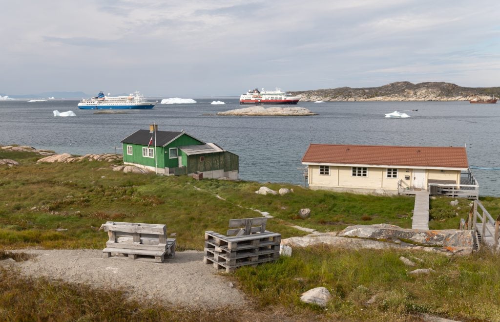Two cottages on the coastline in Illulisat, with two cruise ships in the distance.