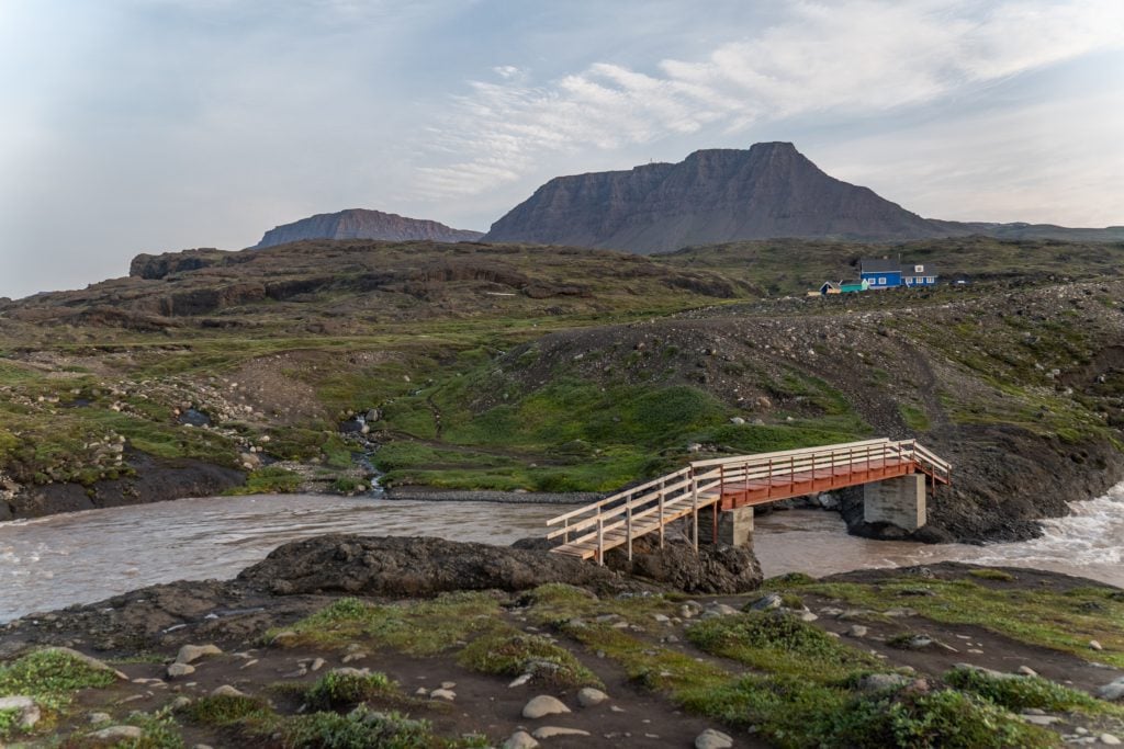 A scene from Disko Island with a wooden bridge crossing a river, a tall, plateau-like mountain and some tiny cottages in the background.