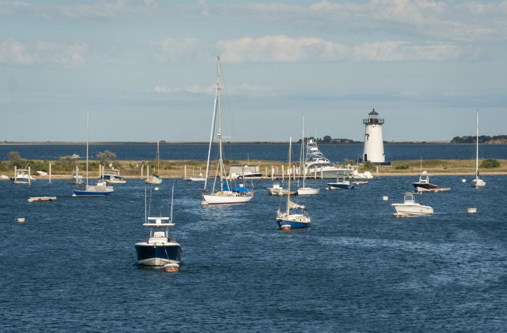 A tiny spit of land topped with a white lighthouse, surrounded by the ocean and topped with lots of small sailboats.