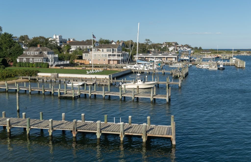 A view above the harbor of wooden docks, sailboats, and perfectly manicured lawns in front of seafront homes.