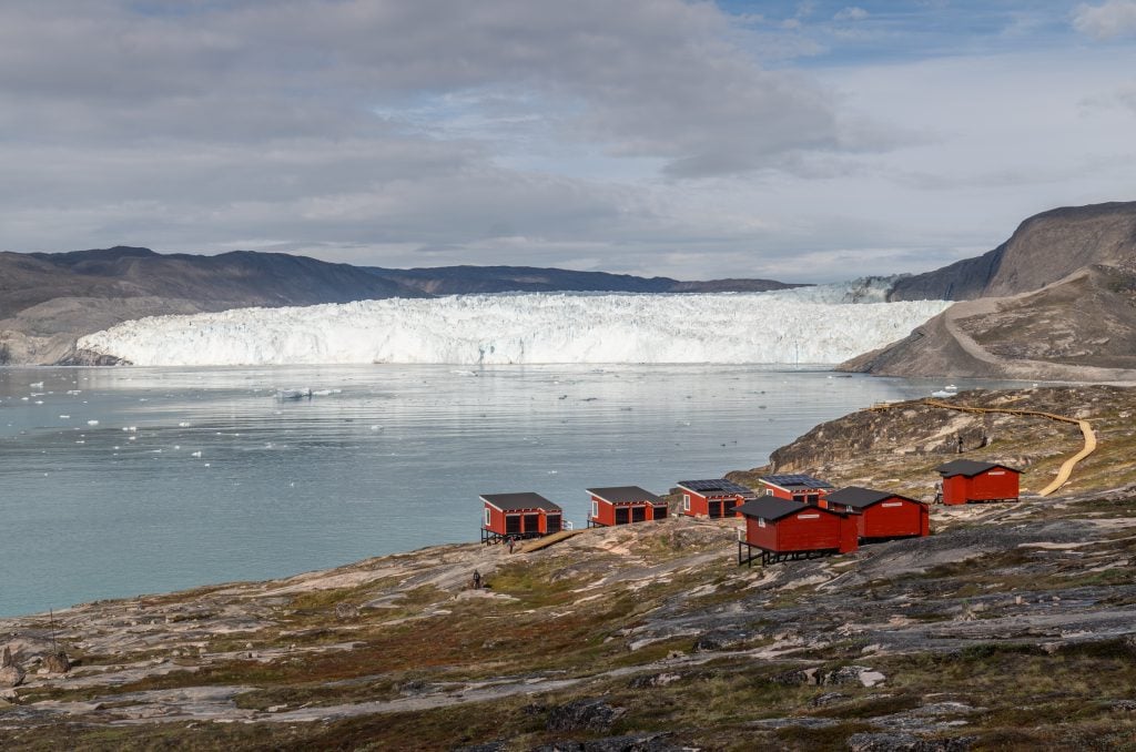 A rocky hill with half a dozen small wooden cabins perched, overlooking a massive glacier in front of a still, pale blue bay.