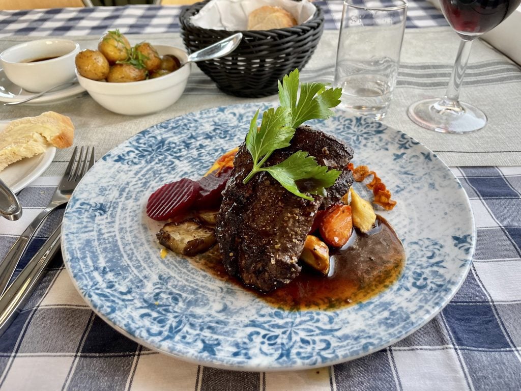 A much more appetizing dish of a reindeer steak and vegetables on a pretty blue and white china plate, a small bowl of new potatoes to the side.