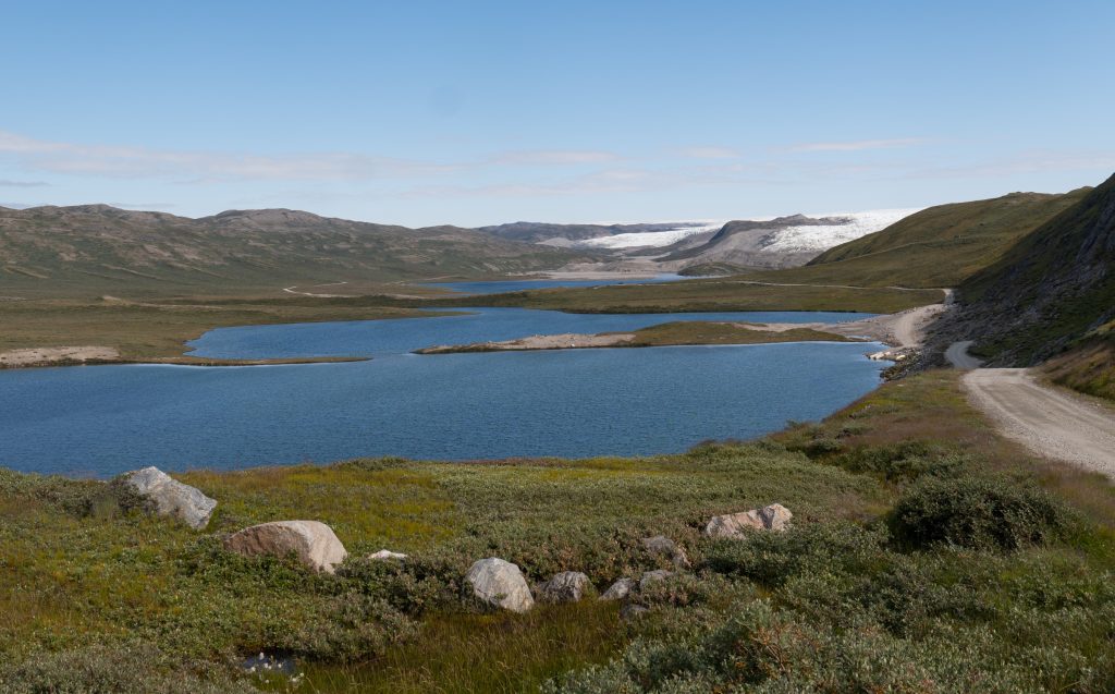 A bright blue lake in Greenland surrounded by green grass. In the distance, you see the white and gray ice cap.