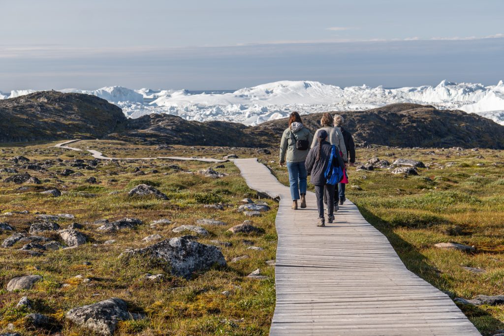 Four women walking down a wooden plank walkway through green scrubby land. In the distance is a giant plate of icebergs.