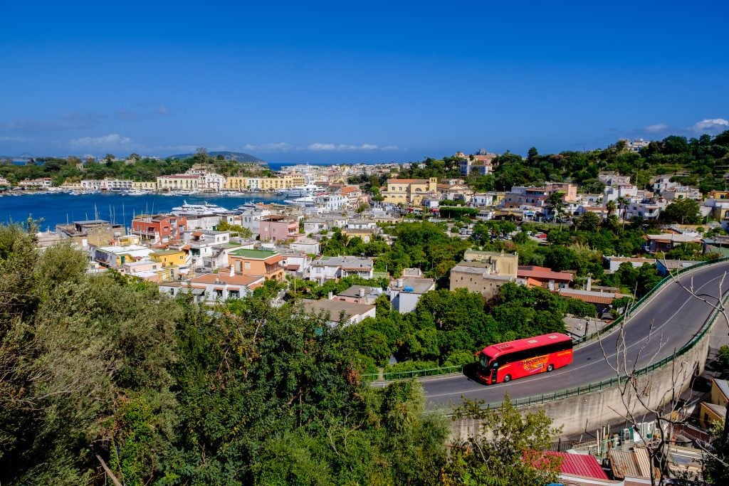 A view of a bright red bus driving through the landscape of Ischia.