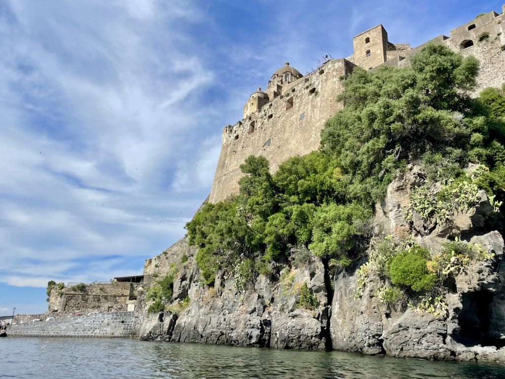 The Castello Aragon's of Ischia, a big stone building rising from the sea, the side covered with green vegetation.