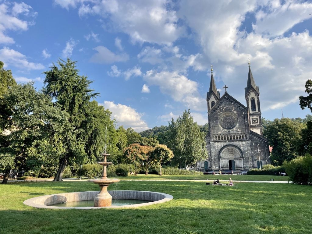 A gothic church set on a big park with a fountain in the middle of the grass.