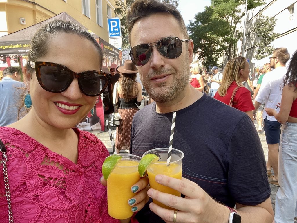 Kate and Charlie taking a smiling selfie in sunglasses, each holding a frozen mango margarita, with a street festival in the background.