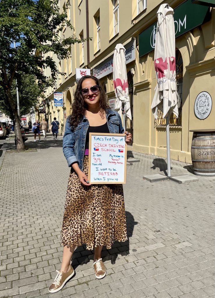 Kate standing holding a sign reading "Kate's first day of Czech Driving School. I am 39 years old. My teacher's name is Honza? Probably Honza. I want to be retired when I grow up."