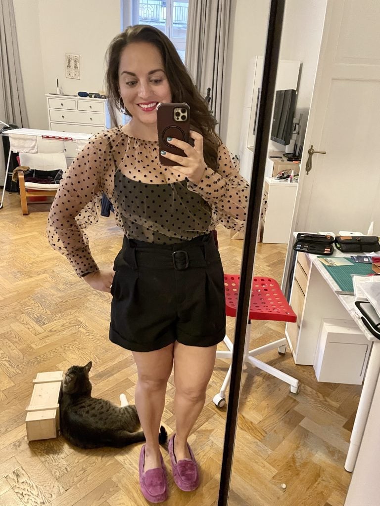 Kate taking a mirror selfie wearing a sheer nude-colored long-sleeved top with tiny black polka dots over a black corset, as well as black shorts. She's inside so wearing her fuzzy purple slippers and Murray the cat hangs out by her feet.