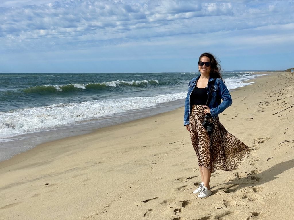 Kate standing on a beach in a leopard print skirt and jean jacket, holding her camera.