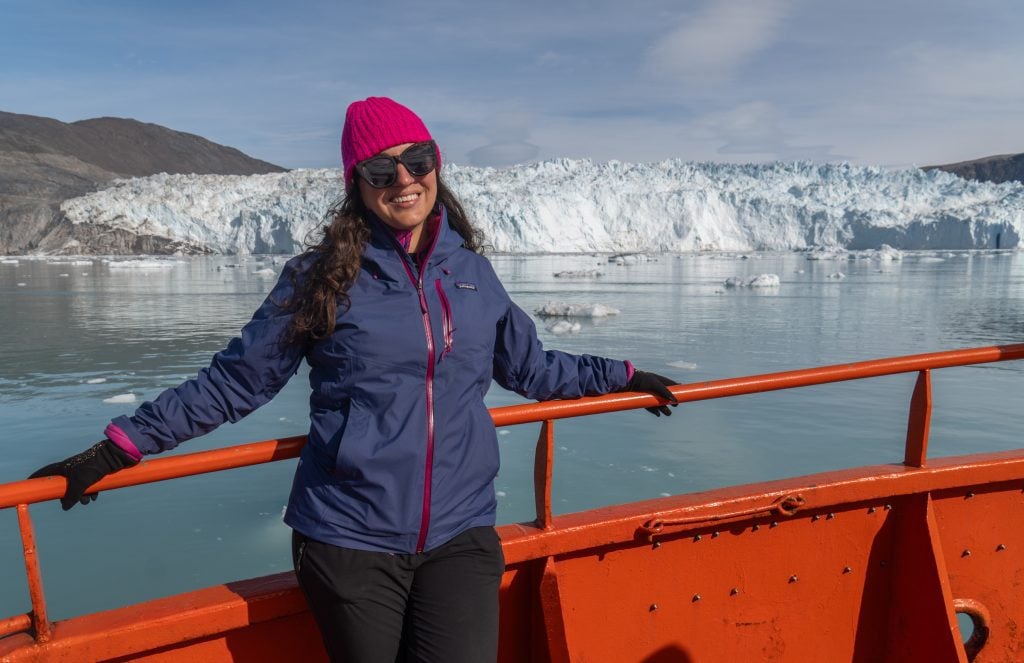 Kate standing on a boat with a big glacier in the background. She wears a purple raincoat, a pink hat, black gloves, and black sunglasses.
