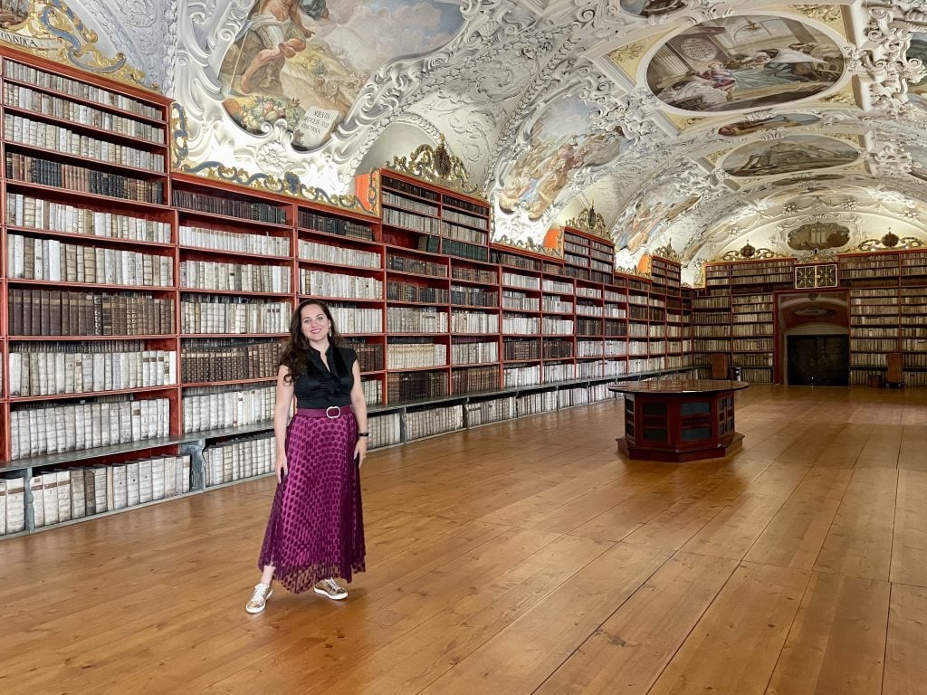 Kate se tient dans une bibliothèque à l'ancienne, au plafond crénelé peint de fresques angéliques. Il y a de vieux livres sur les étagères derrière elle. Elle porte une longue jupe magenta et un haut noir.