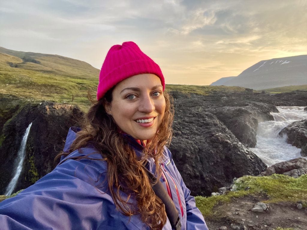 Kate wearing a raincoat and bright pink beanie, standing between two waterfalls at sunrise.