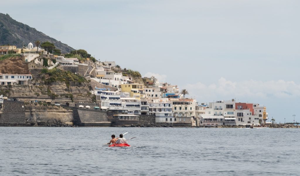 Two people kayaking in a tiny red double kayak in front of a small town in Ischia.