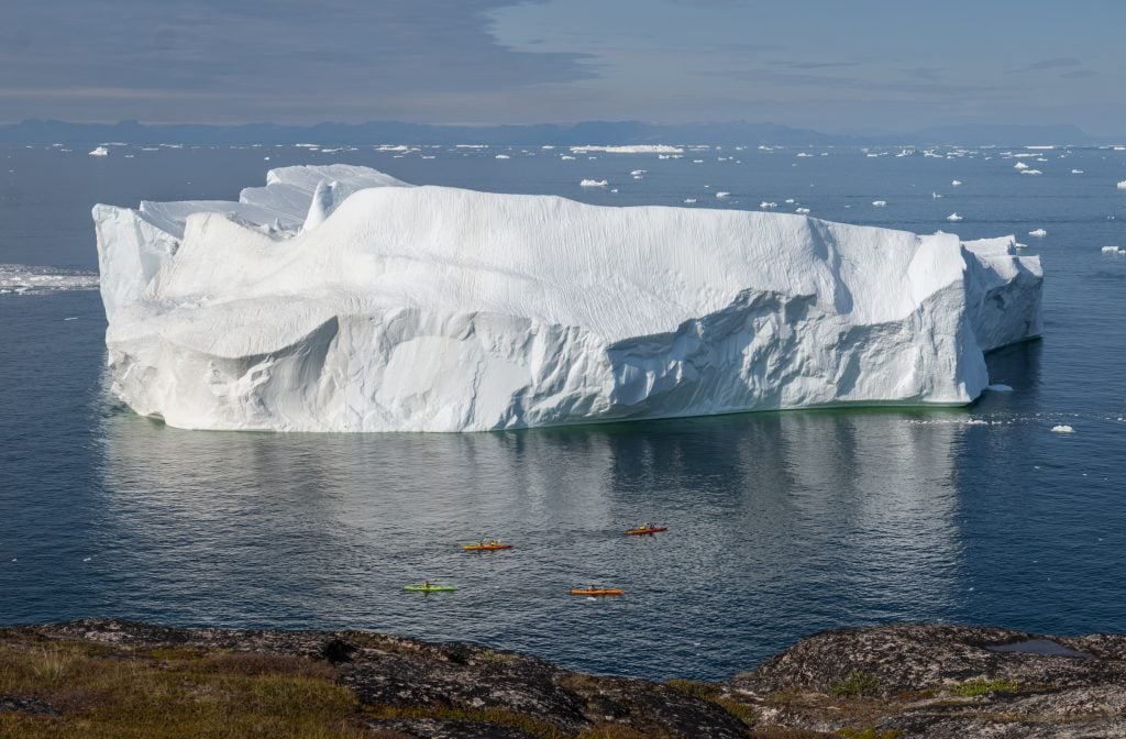 An enormous iceberg in a deep blue bay, with four tiny people in colorful kayaks in the water next to it.