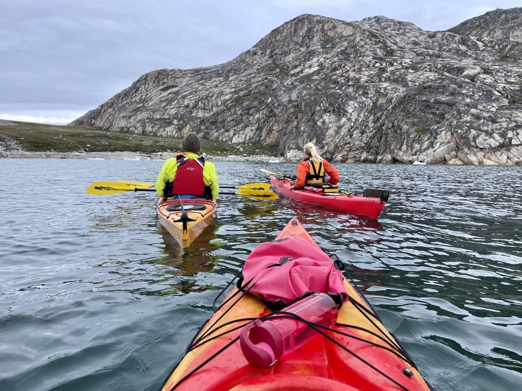Several people kayaking in bright orange and yellow kayaking on a gray bay, surrounded by gray-green rocky landscapes.