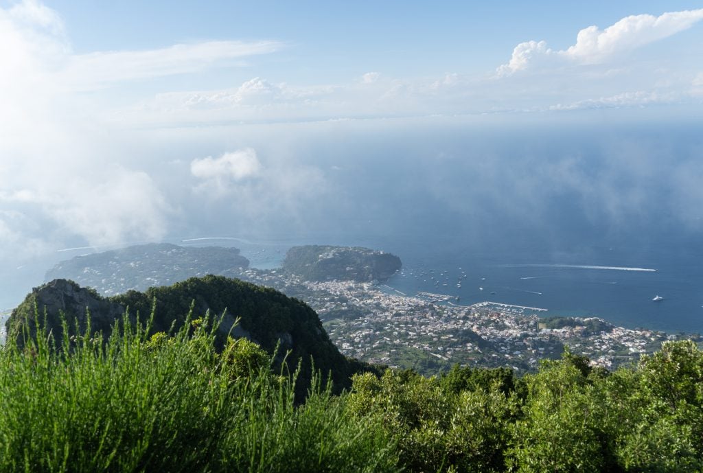 The view from the top of Mount Epomeo in Ischia, with wispy clouds surrounding the landscape and views above small cities squeezed against the coastline. 