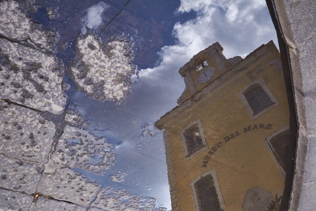 A reflection of the bright yellow Museo del Mare building in a puddle.