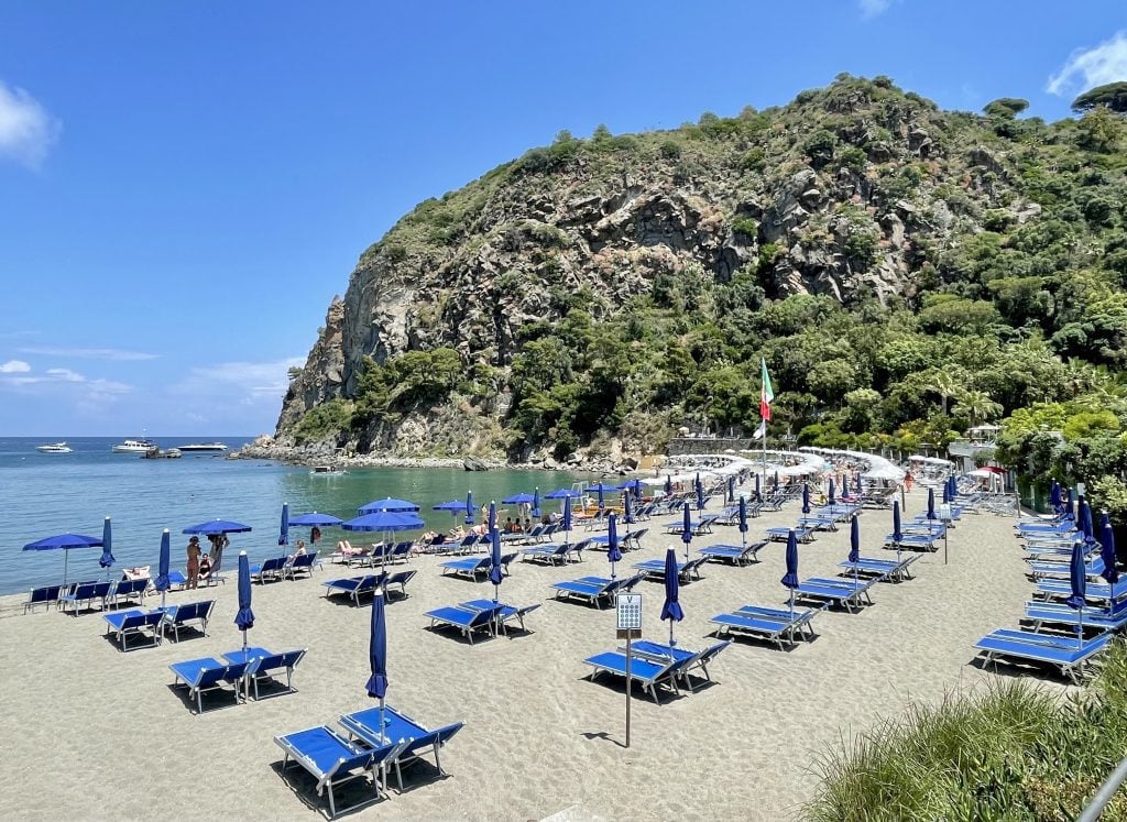A sandy beach covered with bright blue chairs and umbrellas, with a craggy green cliff in the distance.