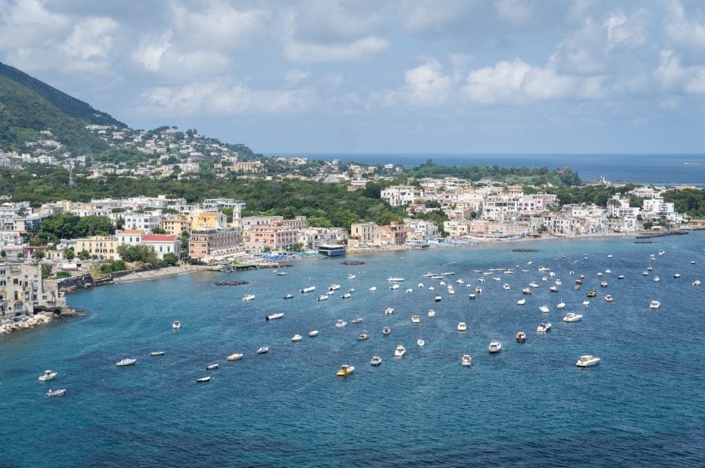 A view from above of the Ischia coastline, with lots of tiny white boats in the water and lots of whitewashed hotels on the water's edge.