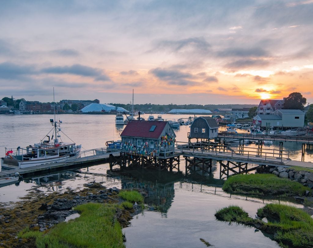 A small Maine island dotted with fishing shacks and piers, underneath a purple and yellow sunrise.