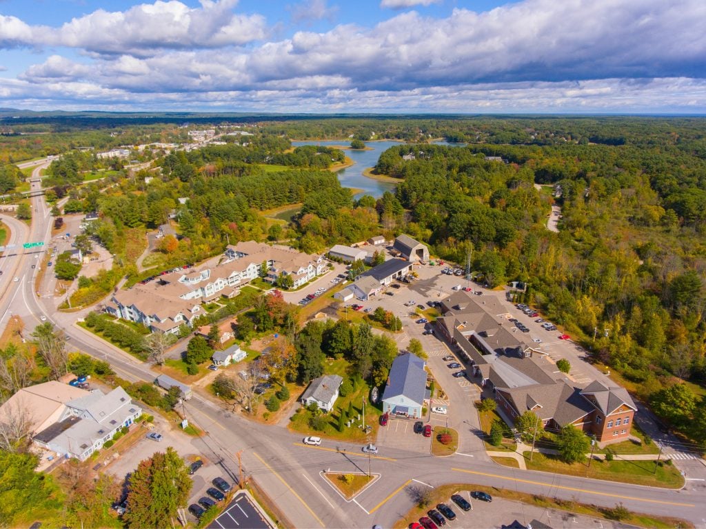 An aerial view of downtown Kittery Maine, with several buildings surrounded by woods and a river in the distance.