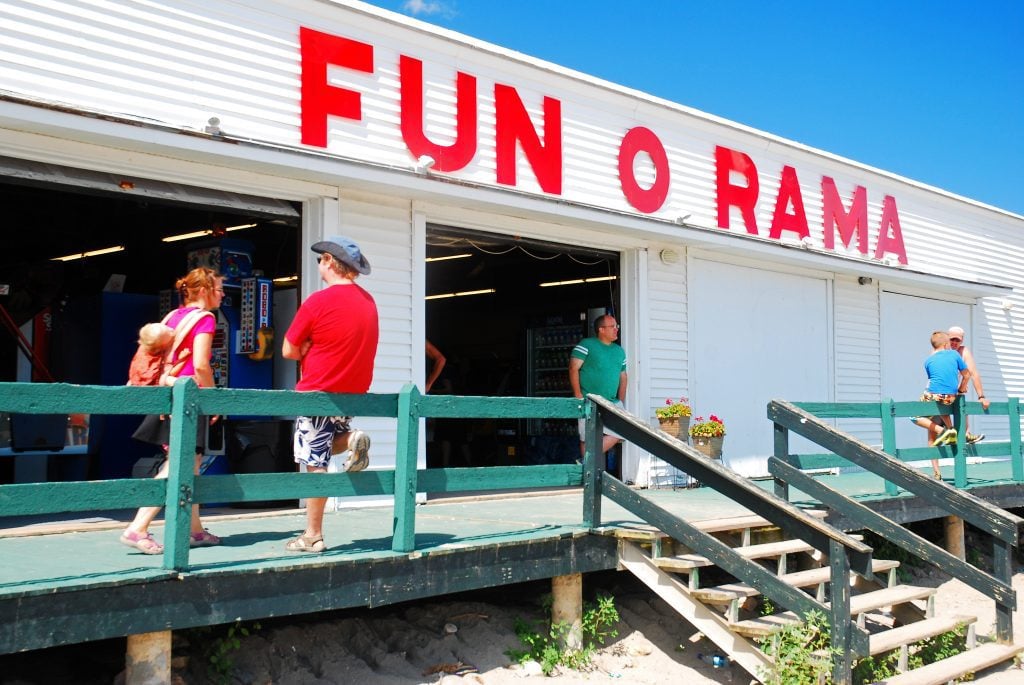 A wooden clapboard building with red letters reading FUN O RAMA on the outside, people hanging out on the porch.