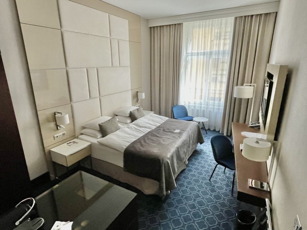 A hotel room with a large gray bed up against a padded wall.