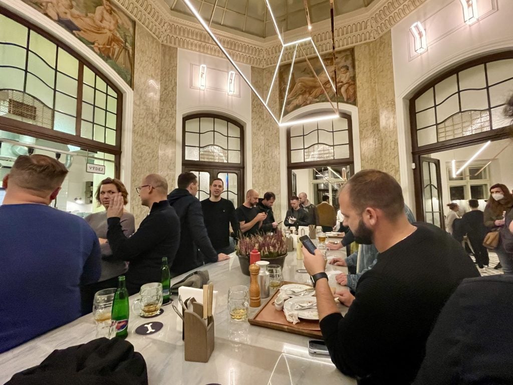 People hanging out and drinking beer around a big marble table in a high-ceilinged pub in Prague.