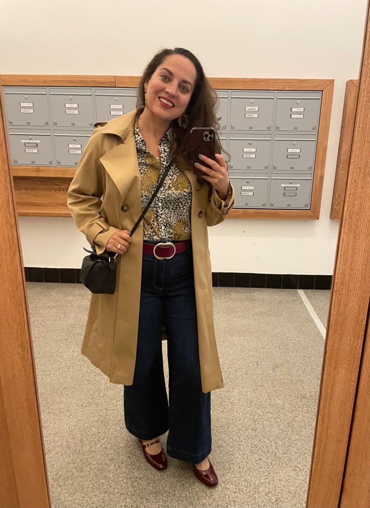 Kate taking a mirror selfie wearing a long beige trench coat, button-down silk shirt with leopards on it, blue wide-leg jeans, purple belt with gold buckle, burgundy patent leather heels, carrying a black square crossbody bag.