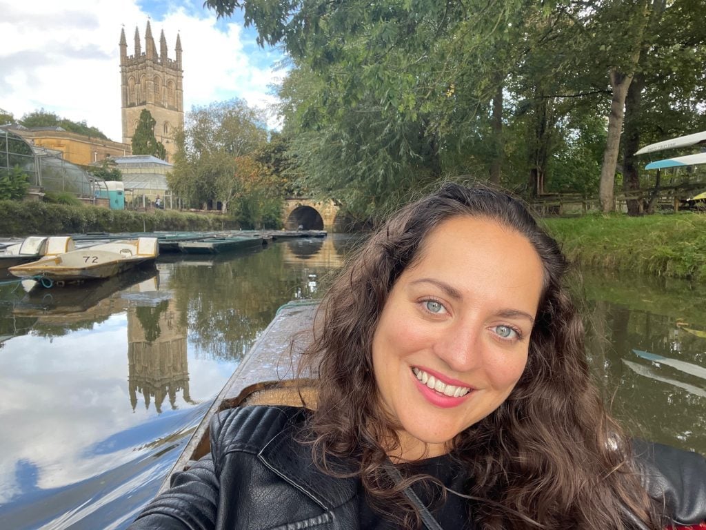 Kate takes a selfie while on a boat in the river in Oxford, a castle tower in the distance behind her.