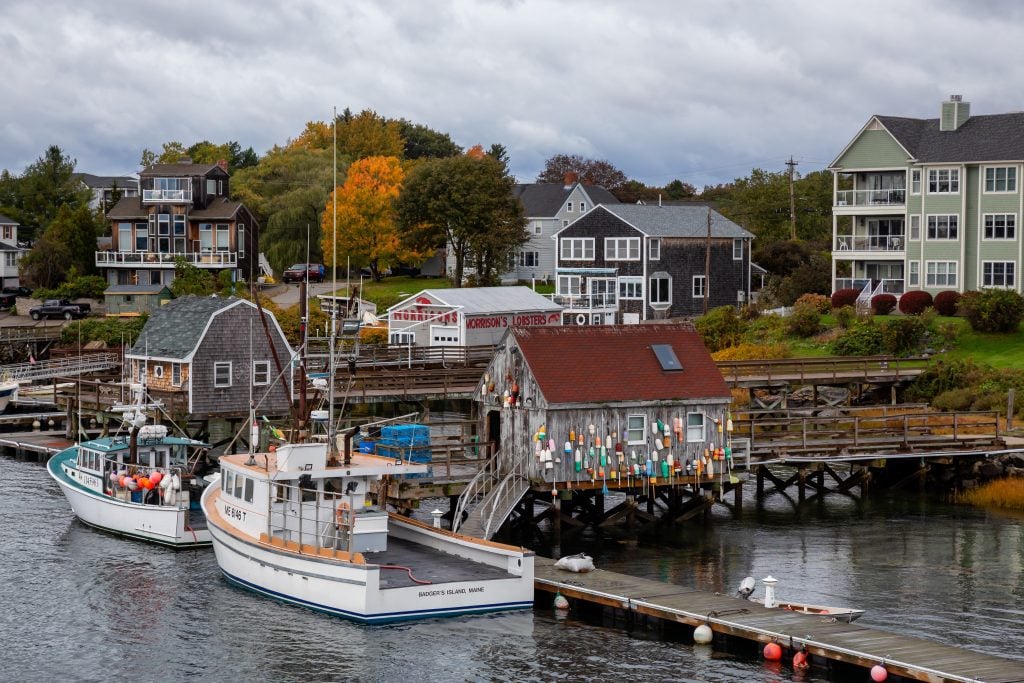 The view of a small town in coastal Maine: fishing shacks covered with colorful buoys and small white boats perched on the edge of a pier, cottages on shore behind them.